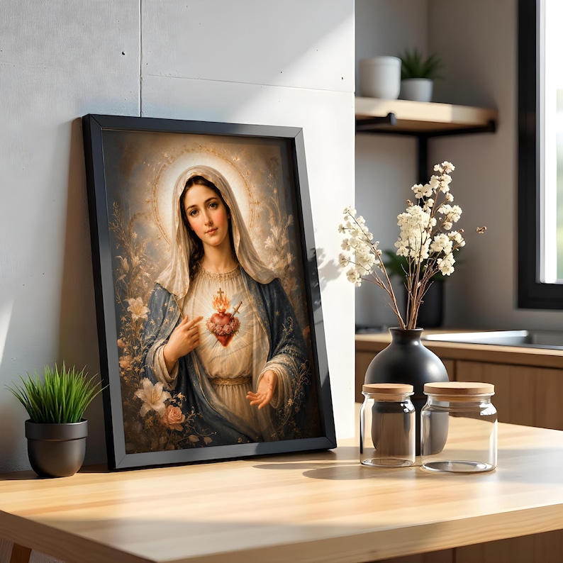 May include: A framed wall art print depicting a figure with a veil and a blue mantle, pointing to a glowing, flaming heart on their chest. The artwork features soft, ethereal lighting, floral accents, and a golden halo, presented in a simple black frame.