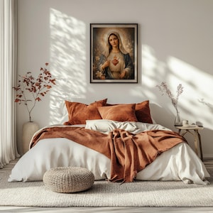 May include: A framed wall art print depicting a woman with a halo and a glowing heart on her chest, centered above a bed with white linens, rust-colored pillows, and a matching throw blanket. A woven pouf sits on a textured rug in the foreground.
