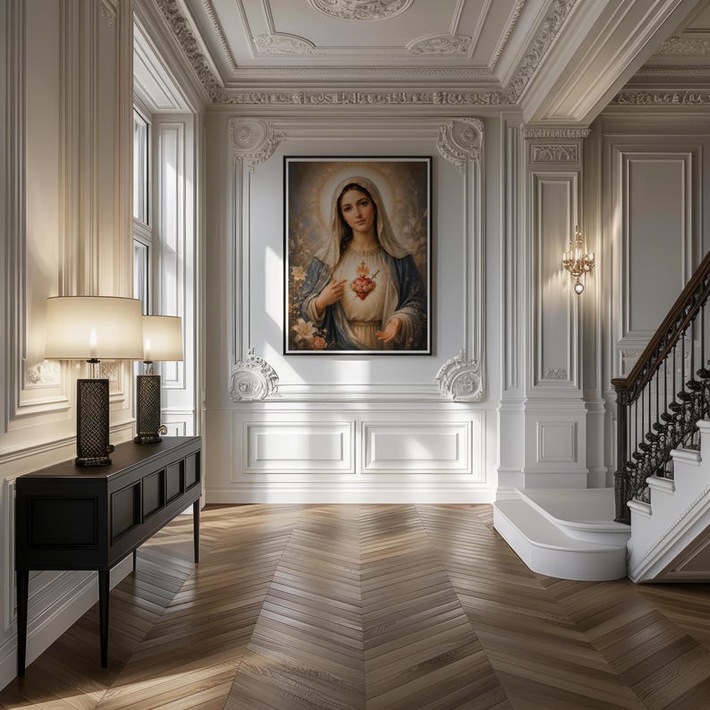 May include: A framed wall art print depicting the Virgin Mary with a glowing Sacred Heart, wearing a blue and white robe, centered on a white paneled wall above a black console table with two lamps in a room with a wooden herringbone floor.