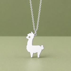 Peut inclure: Un collier argenté avec un pendentif en forme de lama. Le lama est un design simple et stylisé avec un petit œil et une queue touffue. La chaîne délicate est suspendue sur un fond vert.