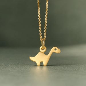 May include: A gold-toned necklace featuring a delicate chain and a small, flat dinosaur-shaped pendant. The pendant has a smooth, matte finish and is suspended from a simple circular jump ring, showcasing a minimalist silhouette of a long-necked creature.