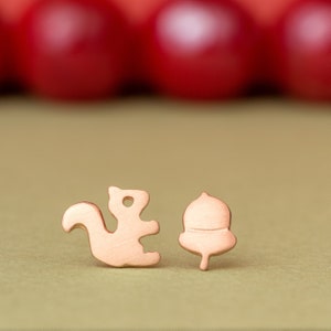 May include: A pair of gold-toned earrings featuring a squirrel and an acorn design. The earrings are on a green surface.