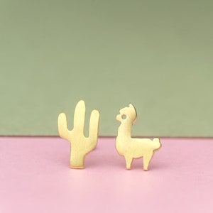May include: Gold-colored stud earrings featuring a cactus and a llama. The earrings are set against a pink and green background. The cactus has a three-pronged design, and the llama is standing.