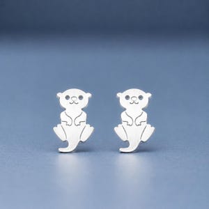 May include: A pair of silver-toned stud earrings shaped like small otters. Each earring features a brushed metal finish with engraved lines defining the otter's face, paws, and tail, creating a symmetrical design of the animal in a seated position.