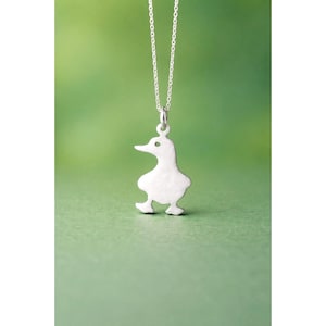 Op de afbeelding: Een zilverkleurige ketting met een eendenhanger. De hanger is een eenvoudige, gestileerde eendvorm met een klein oogdetail. De ketting is delicaat en zilverkleurig, tegen een zachte groene achtergrond.