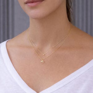 May include: A delicate gold necklace set featuring two strands. The top strand has a small bead, while the lower strand displays a Virgo zodiac symbol. The necklaces are worn on a person wearing a white shirt.