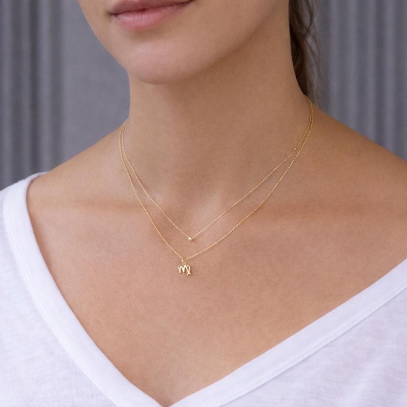 May include: A delicate gold necklace set featuring two strands. The top strand has a small bead, while the lower strand displays a Virgo zodiac symbol. The necklaces are worn on a person wearing a white shirt.