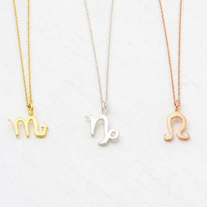 May include: Three necklaces with zodiac symbols. One is gold with a Scorpio symbol, one is silver with a Capricorn symbol, and one is rose gold with a Leo symbol. Each necklace has a delicate chain.