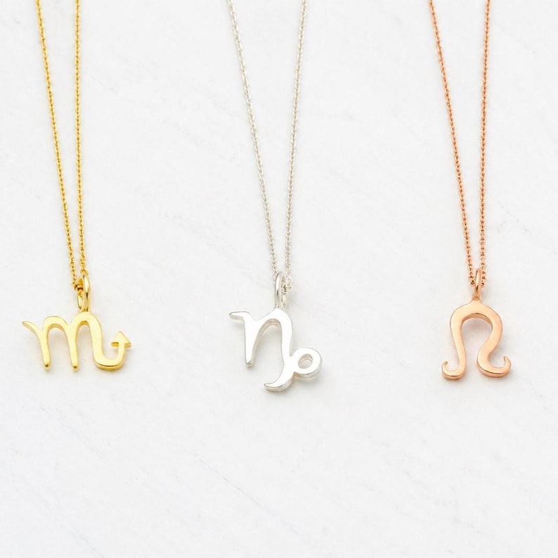 May include: Three necklaces with zodiac symbols. One is gold with a Scorpio symbol, one is silver with a Capricorn symbol, and one is rose gold with a Leo symbol. Each necklace has a delicate chain.