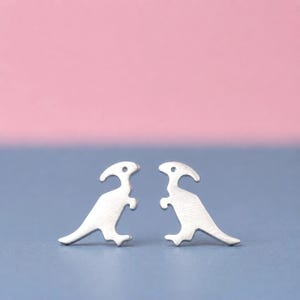 Puede incluir: Un par de pendientes de botón de metal plateado con forma de dinosaurios Parasaurolophus. Cada pendiente presenta un acabado mate liso con un pequeño recorte circular que representa el ojo, orientados uno frente al otro en diseño espejo.