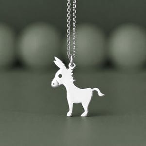 Könnte beinhalten: Halskette mit einem silberfarbenen Esel-Anhänger. Der Anhänger ist eine stilisierte Silhouette eines Esels mit kleinem Schwanz und langen Ohren. Die Halskette ist ebenfalls silberfarben.