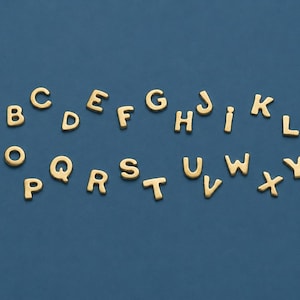 Op de afbeelding: Goudkleurige alfabetletters op een donkerblauwe achtergrond. De letters zijn in een schreeflettertype en zijn verspreid over het oppervlak. De letters zijn in hoofdletters en kleine letters.