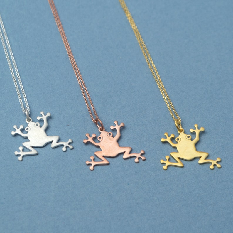May include: Three silver frog necklaces on a blue background. The necklaces are in silver, rose gold, and gold.