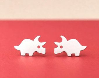 May include: A pair of silver-toned stud earrings shaped like triceratops dinosaurs. Each earring features a smooth, flat surface with a cutout circle representing the eye, showcasing a clean silhouette of the prehistoric creature in profile.