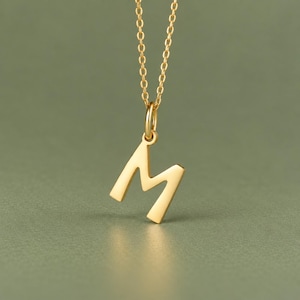 May include: A gold-toned necklace featuring a capital letter M pendant suspended from a delicate matching chain. The pendant has a smooth, polished finish and is attached to the chain by a small circular jump ring, creating a simple design.