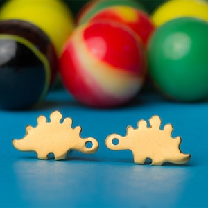 Könnte beinhalten: Zwei goldfarbene Metall-Dinosaurier-Anhänger, jeder mit einem kleinen Loch zum Anbringen an einer Halskette oder einem Armband. Die Anhänger sind auf einer blauen Oberfläche einander zugewandt.