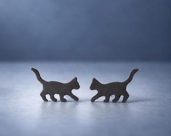 Op de afbeelding: Een paar donkergrijze metalen oorknopjes in de vorm van lopende katten. Elke oorbel toont het silhouet van een kat met een omhoogstaande staart, puntige oren en vier poten. De oorbellen zijn naast elkaar op een plat oppervlak geplaatst.
