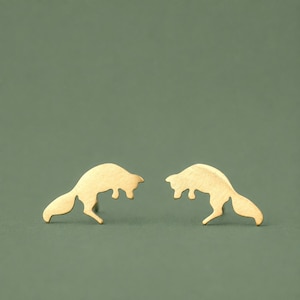 May include: A pair of gold-toned stud earrings shaped like leaping foxes. Each earring features a smooth, matte metallic finish and a silhouette of a fox in a mid-air jumping pose with its tail extended behind it and legs tucked underneath.