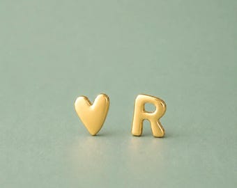 Op de afbeelding: Goudkleurige oorstekers. Eén oorbel heeft de vorm van een hart en de andere de letter "R". De oorbellen staan op een lichtgroene achtergrond.
