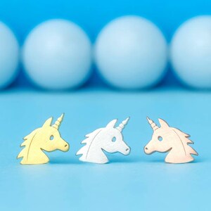 Könnte beinhalten: Drei Silhouetten von Einhornköpfen in Gold, Silber und Roségold auf blauem Hintergrund.