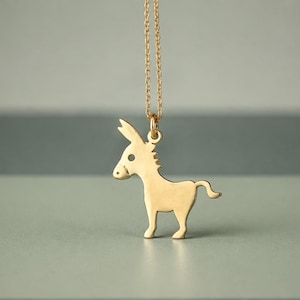May include: A gold-toned necklace featuring a flat, silhouette-style pendant shaped like a donkey. The pendant is suspended from a delicate, thin gold-toned chain. The donkey design includes a small circular cutout for the eye and a curved tail.