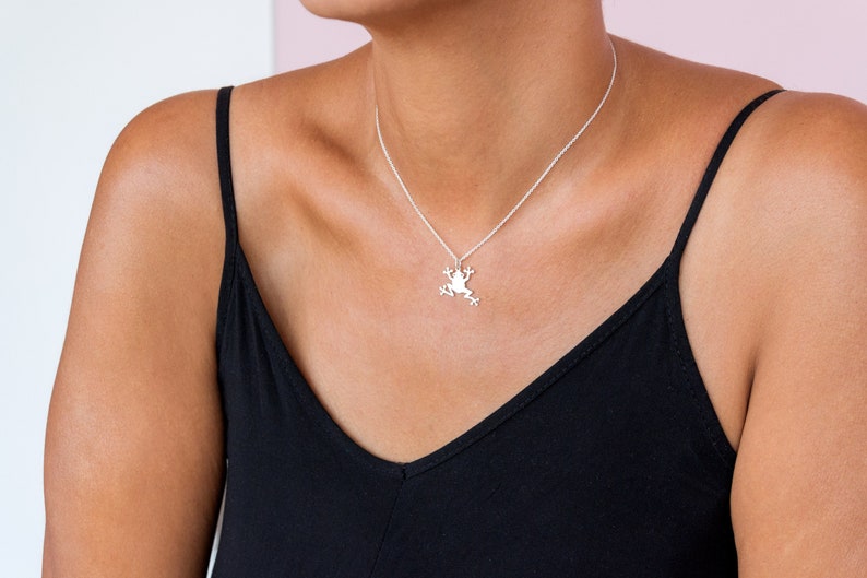 May include: A silver necklace with a frog pendant. The necklace is worn by a person with a black tank top.