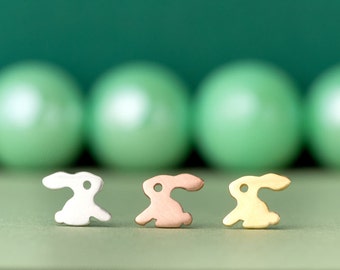 May include: Three small rabbit figurines made of metal. The rabbits are silver, copper, and gold in colour. The rabbits are sitting on a green surface.