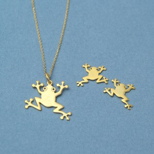 May include: A gold necklace with a frog pendant and two smaller frog charms. The frogs are all facing the same direction with their legs outstretched.