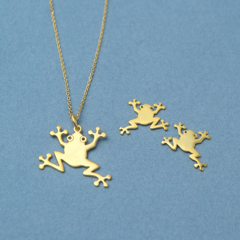 May include: A gold necklace with a frog pendant and two smaller frog charms. The frogs are all facing the same direction with their legs outstretched.