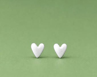 May include: A pair of small, silver-toned heart-shaped stud earrings. Each earring features a smooth, matte finish and a simple, symmetrical heart silhouette, designed to be worn as a matching set on the earlobes for a clean, understated look.