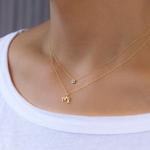 May include: Two delicate gold necklaces. The top necklace features a small, round charm with a blue center. The bottom necklace has a charm in the shape of the Virgo zodiac symbol. The necklaces are worn against a white shirt.