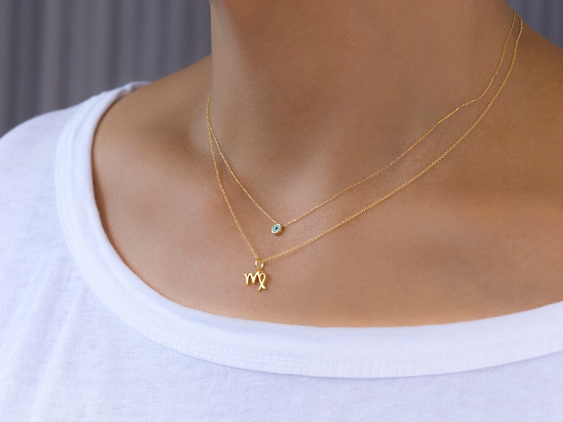 May include: Two delicate gold necklaces. The top necklace features a small, round charm with a blue center. The bottom necklace has a charm in the shape of the Virgo zodiac symbol. The necklaces are worn against a white shirt.