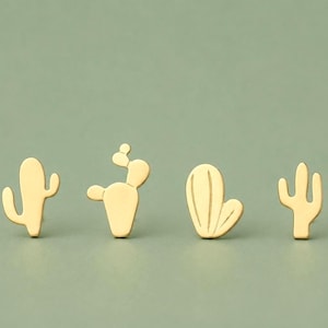 Op de afbeelding: Vier goudkleurige metalen oorknopjes, elk in de vorm van een andere cactus. De ontwerpen omvatten een hoge saguaro, een vijgcactus met een klein rond schijfje, een ronde toncactus met gegraveerde lijnen en een slanke cactus met meerdere armen.