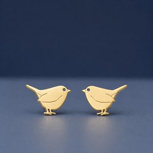 Pode incluir: Um par de brincos de botão dourados em forma de pássaro. Cada brinco apresenta um pequeno olho redondo e um corpo curvo com cauda e pés. Os brincos são exibidos em um fundo azul escuro.
