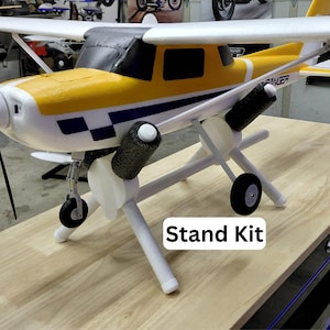 May include: A model airplane with a yellow and white fuselage, black propeller, and white wings. The plane is on a white stand with the text "Stand Kit". The plane has a black and white checkered pattern on the side.