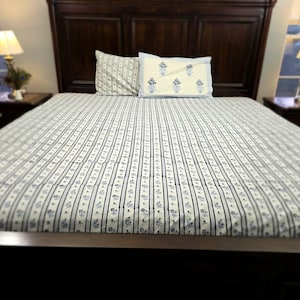 May include: A white bed with a blue and white striped bedspread. The bed has a wooden headboard and footboard. There are two pillows on the bed, one with a blue and white floral pattern and one with a solid blue color.
