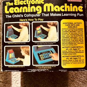 May include: The Coleco Electronic Learning Machine is a blue and yellow toy computer with a screen, buttons, and a slot for a cartridge. The box shows a child using the machine and includes information about additional activity card sets.