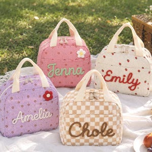 Puede incluir: Cuatro bolsas de almuerzo rectangulares con asas superiores y cierres de cremallera. Las bolsas presentan nombres bordados: Jenna en una bolsa floral rosa, Emily en una bolsa blanca de fresas, Amelia en una bolsa floral morada y Chole en una bolsa a cuadros beige.