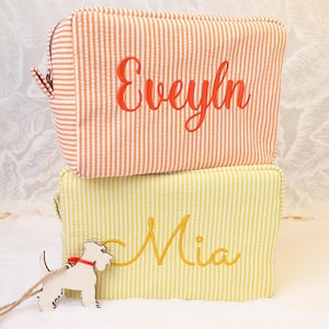 May include: Two seersucker cosmetic bags, one orange and white striped, the other yellow and white striped. The orange bag has the name "Evelyn" embroidered in orange. The yellow bag has the name "Mia" embroidered in yellow. A wooden dog tag is attached.