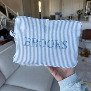 May include: A rectangular zippered pouch made of light blue and white seersucker fabric with vertical stripes. The name BROOKS is embroidered in a matching light blue thread across the center of the front panel of the pouch.
