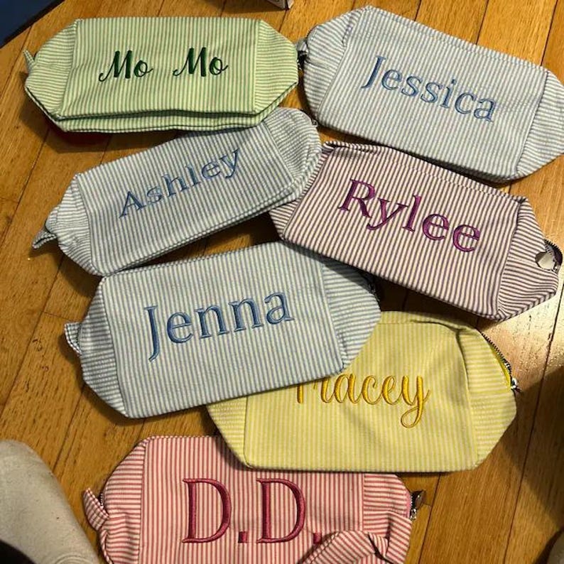 May include: A collection of seven rectangular zippered pouches made of striped fabric in various colors including light green, light blue, purple, yellow, and pink. Each pouch features a name embroidered in a contrasting thread color on the front.