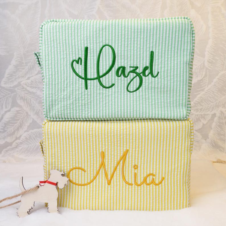 May include: Two rectangular zippered pouches made of seersucker fabric. The top pouch is light green and white striped with the name Hazel embroidered in dark green. The bottom pouch is yellow and white striped with the name Mia embroidered in yellow.