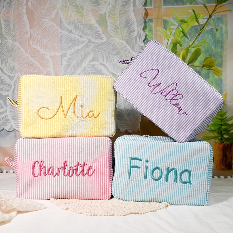 May include: Four rectangular zippered pouches made of seersucker fabric with white stripes. The yellow pouch features the name Mia, the pink pouch says Charlotte, the light blue pouch says Fiona, and the purple pouch is embroidered with Willow.