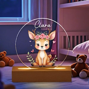 May include: A decorative night light with a cartoon deer wearing a floral crown, butterflies, and the name "Clara". The deer is set within a circular frame, lit by a warm light from a wooden base.