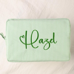 May include: A rectangular zippered pouch featuring a light green and white vertical pinstripe pattern. The front of the pouch is embroidered with the name Hazel in a dark green cursive script, with a small heart integrated into the first letter.