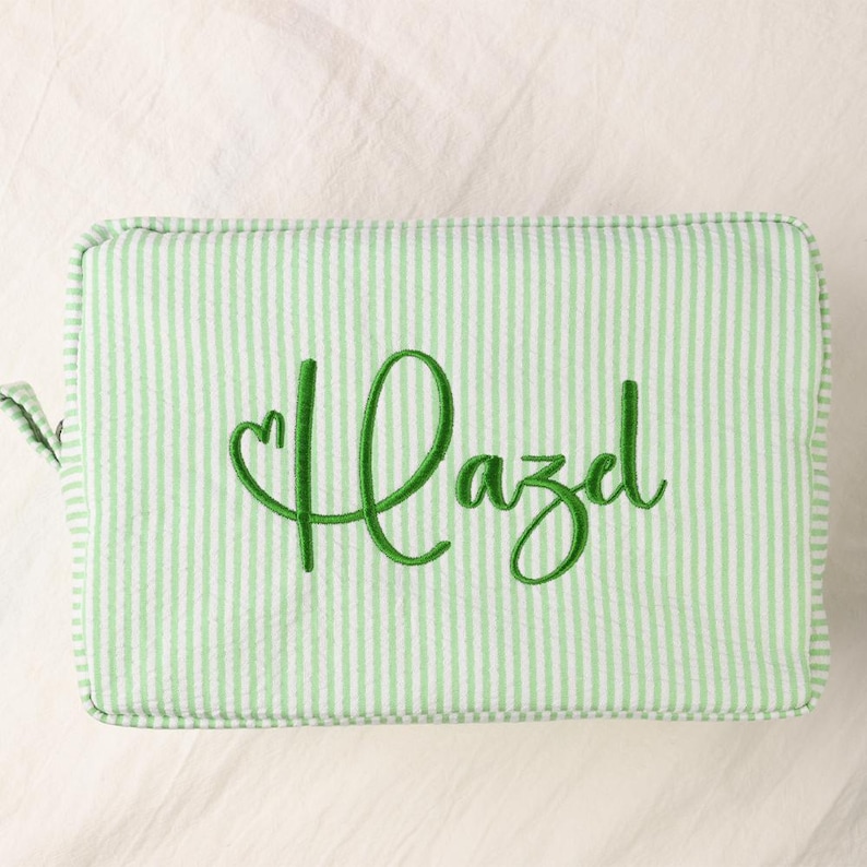 May include: A rectangular zippered pouch featuring a light green and white vertical pinstripe pattern. The front of the pouch is embroidered with the name Hazel in a dark green cursive script, with a small heart integrated into the first letter.