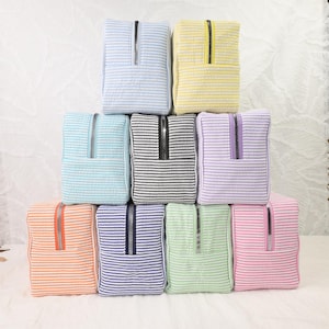 May include: A collection of ten rectangular zippered pouches arranged in a pyramid stack. Each pouch features a seersucker texture with horizontal white stripes alternating with colors including light blue, yellow, teal, black, lavender, orange, purple, green, and pink.