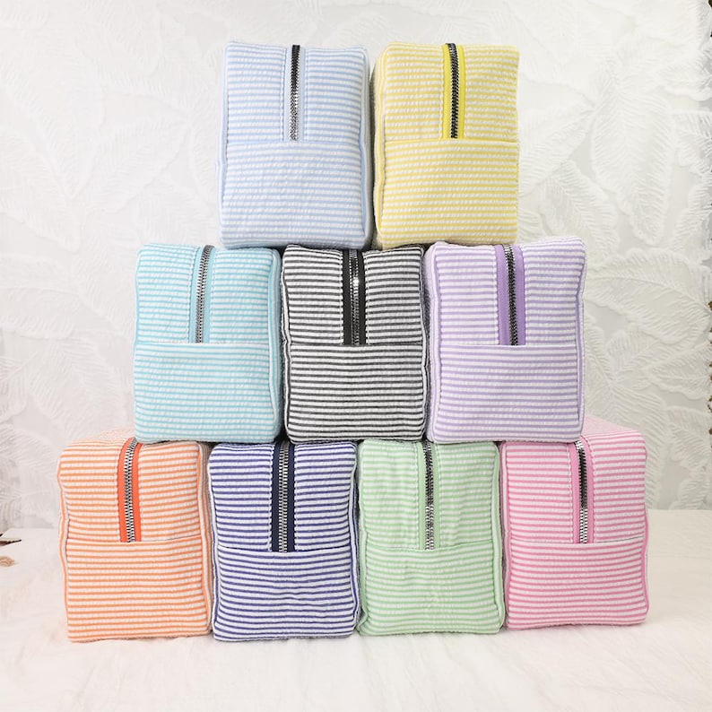 May include: A collection of ten rectangular zippered pouches arranged in a pyramid stack. Each pouch features a seersucker texture with horizontal white stripes alternating with colors including light blue, yellow, teal, black, lavender, orange, purple, green, and pink.