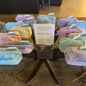 May include: A collection of striped zippered pouches in various pastel colors, each featuring an embroidered name. A central white sign displays the text "Behind every successful woman is a tribe of other successful women who have her back" and "dinnerdesigners".