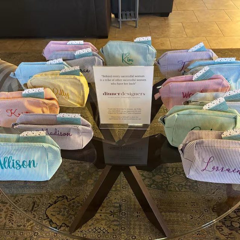 May include: A collection of striped zippered pouches in various pastel colors, each featuring an embroidered name. A central white sign displays the text "Behind every successful woman is a tribe of other successful women who have her back" and "dinnerdesigners".